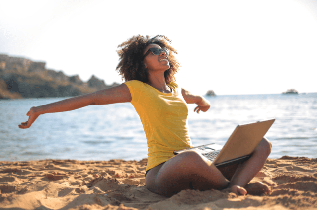Woman with arms outstretched sitting on the beach working remotely with laptop